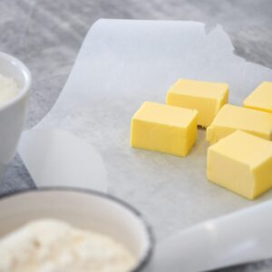 Close-up of butter cubes on parchment paper with bowls of ingredients in a kitchen setting.