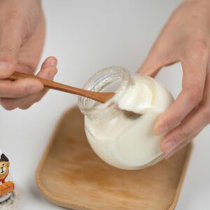 Close-up of hands serving creamy yogurt from a jar using a wooden spoon, with a cute figurine on the side.